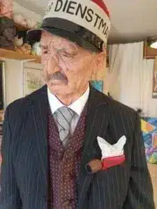 An elderly gentleman in a pinstripe suit and tie wears a hat with German lettering, has a cigar and handkerchief in his breast pocket and stands in the house with a serious expression.