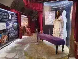 A museum exhibition with information boards, a mannequin in a white dress, a purple bench and decorated chairs in a room with red curtains and a stone floor.