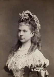 A vintage portrait of a young woman in an elaborate 19th century dress with lace details and flowers in her curly hair, posing in profile against a plain background.