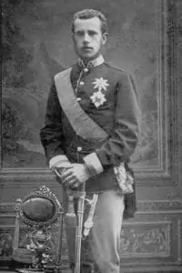 Un joven con uniforme militar formal, medallas y fajín, de pie junto a una silla ornamentada, sostiene una espada en la mano. Se muestra una foto antigua en blanco y negro con fondo pintado.