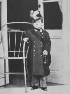 A small child stands next to a large metal chair, wears an oversized coat and a hat with decorations and poses inside in front of a door.