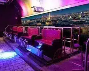 In an indoor entertainment venue, there are seats for rides with red upholstery and black trim in front of a mural with a cityscape under colorful lighting.
