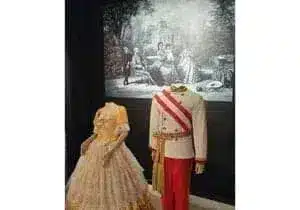 Two headless mannequins in period clothing - an ornate yellow dress and a white military uniform with a red sash - stand in front of a black and white vintage painting depicting aristocrats.