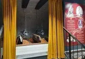 A small room with golden curtains drawn to one side reveals two vintage-style wooden electric chairs under hanging metal head covers. On the wall to the right is an information board with text and pictures.