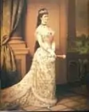 豪華な白いロングドレスを着た女性が、カーテンのかかった窓の横に立っている。茶色の長い髪にティアラをつけ、手には扇を持っている。植物を背景に、フォーマルなフレームの中でエレガントにポーズをとっている。