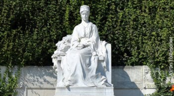Weiße Marmorstatue einer sitzenden Frau in wallendem Gewand mit gefalteten Händen vor einem Hintergrund aus grünem Efeu. Die Statue ruht auf einem Steinsockel mit geschnitzten Details.