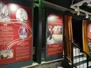 Large red information boards with historical pictures and texts are mounted on a wall next to a staircase with a black banister and golden curtains in a museum or exhibition.