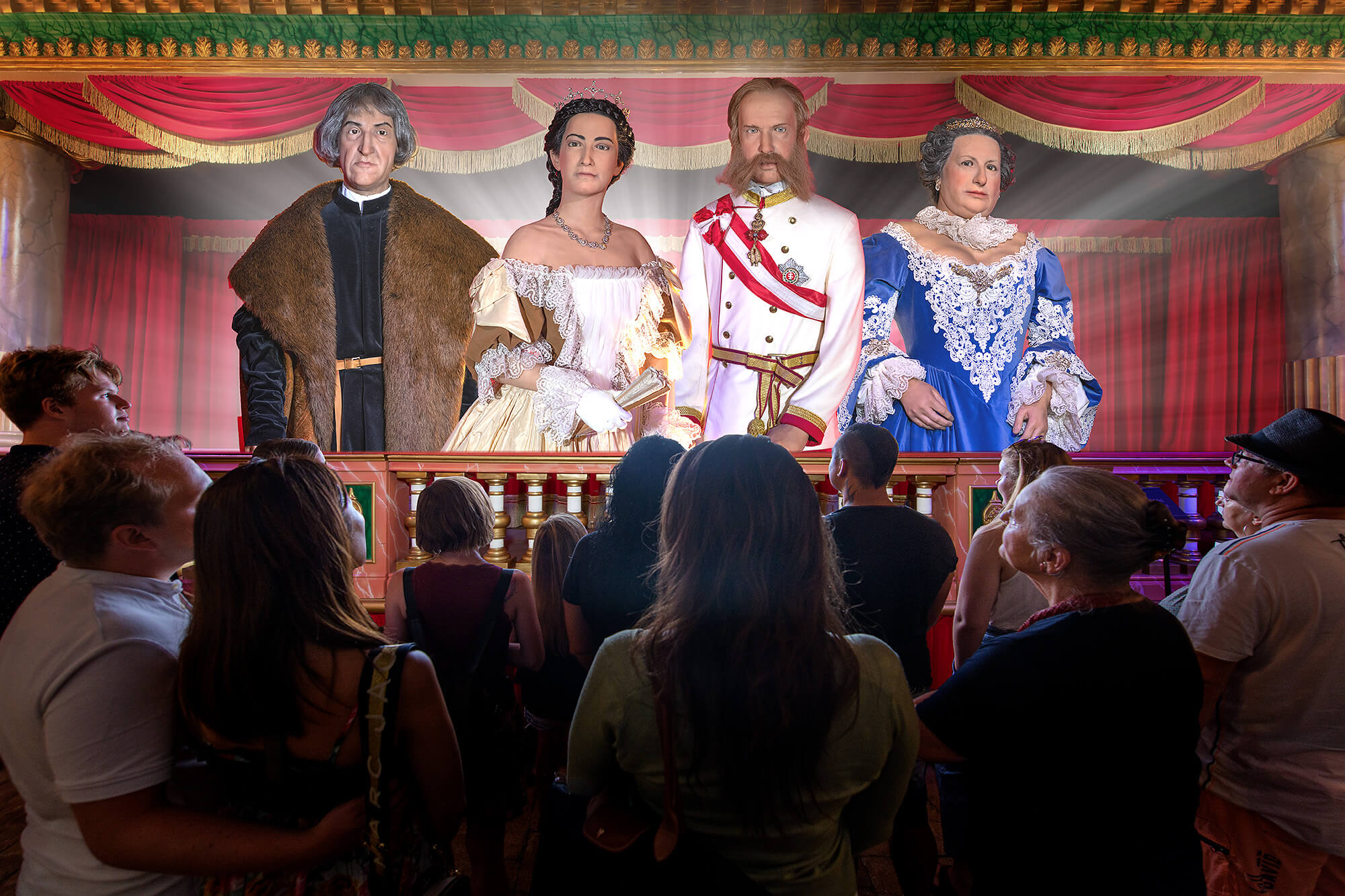 A group of people in a theater look at large, illuminated portraits of four historical figures in royal dress displayed on the stage, with red curtains and ornate decor in the background.
