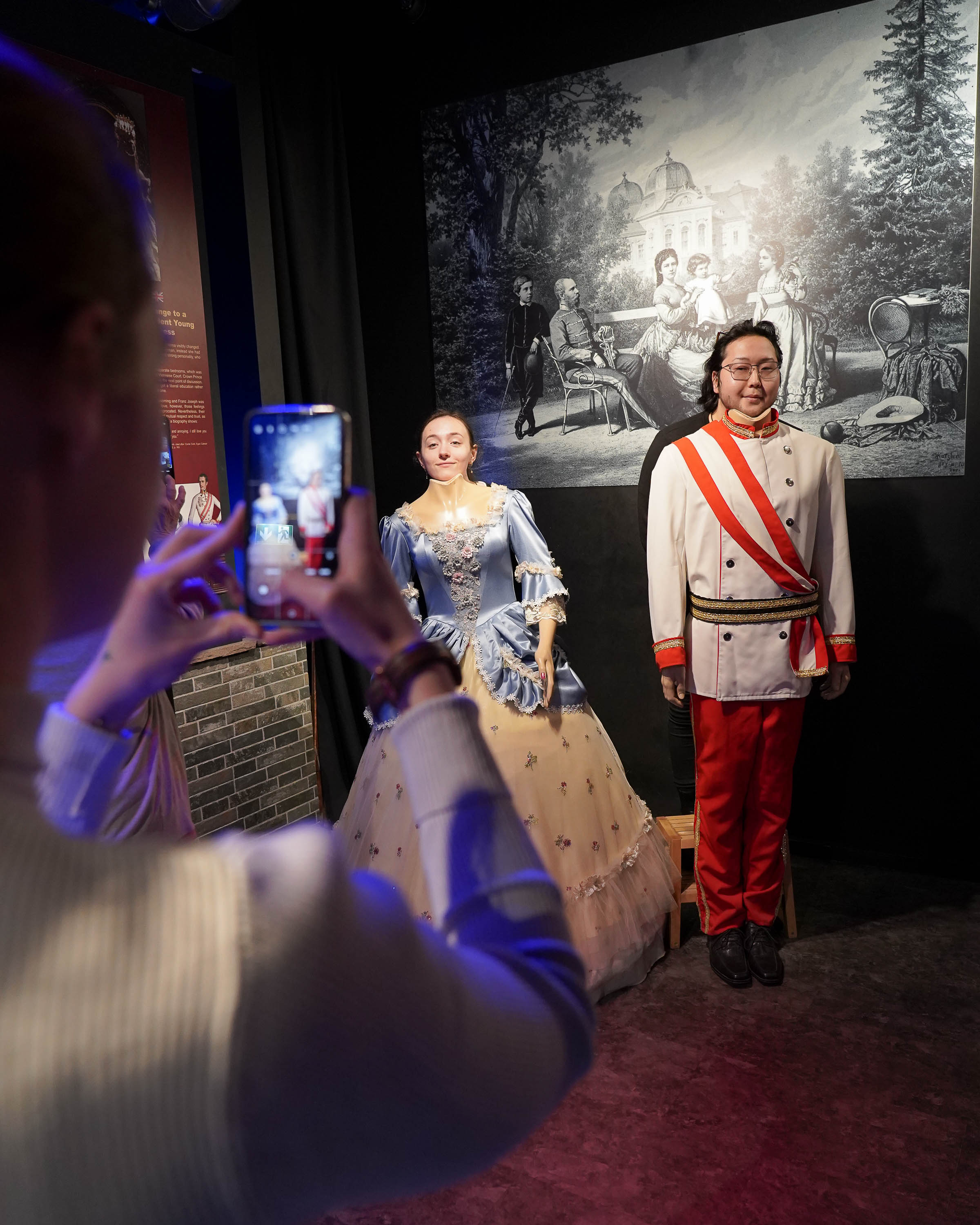 A person uses a phone to take a photo of two wax figures dressed in elaborate 18th or 19th century European costumes, posing in front of an old-fashioned black and white background.  