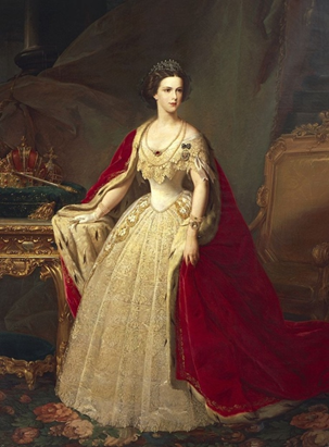 Eine Frau in einem verzierten cremefarbenen Kleid mit goldenen Details und einem roten Samtumhang steht in einem reich geschmückten Raum neben einem vergoldeten Tisch und einer Krone, trägt eine Tiara und Schmuck.