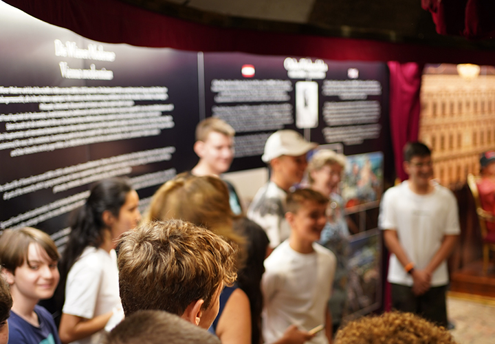 Eine Gruppe junger Menschen steht in einem Innenraum zusammen, lächelt und blickt auf eine museumsähnliche Ausstellung mit Texttafeln und Bildern im Hintergrund. Die Szene wirkt lebendig und ungezwungen.