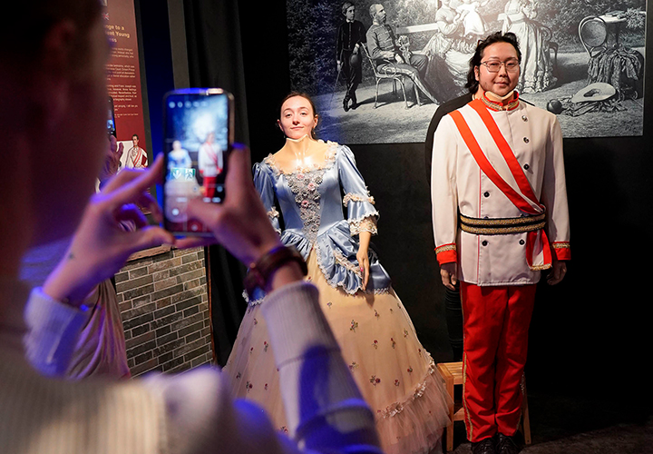 Eine Person macht ein Foto von zwei Personen in historischen Kostümen; die eine trägt ein blau-weißes Kleid, die andere eine weiße Militäruniform mit einer roten Schärpe. Sie posieren vor einer schwarz-weißen historischen Kulisse.