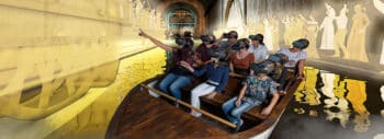 Eine Gruppe von Personen mit Virtual-Reality-Headsets sitzt in einem Holzboot auf dem Wasser, umgeben von digitalen Bildern antiker Figuren und Strukturen, die auf die Wände projiziert werden.