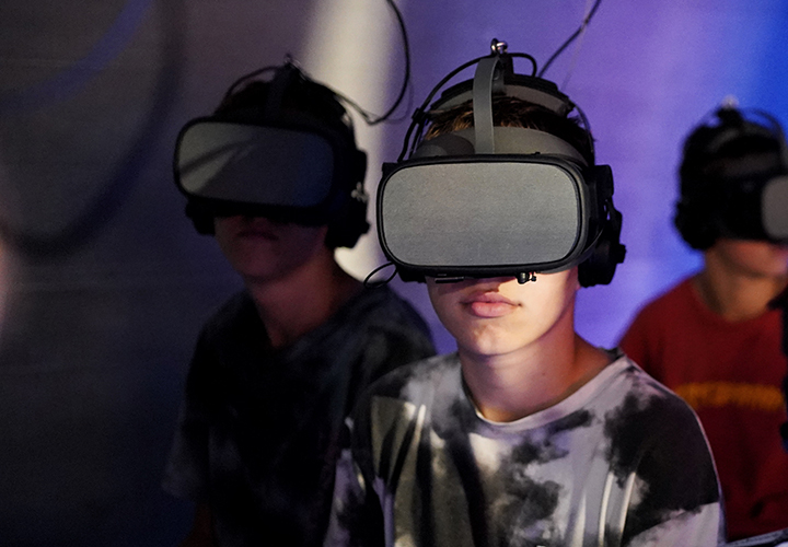 Drei junge Menschen, die Virtual-Reality-Headsets tragen, sitzen in einem Innenraum, der teilweise von bunten Lichtern beleuchtet wird. Der Fokus liegt auf der Person im Vordergrund, die ein schwarz-weißes Hemd mit Krawattenmuster trägt.