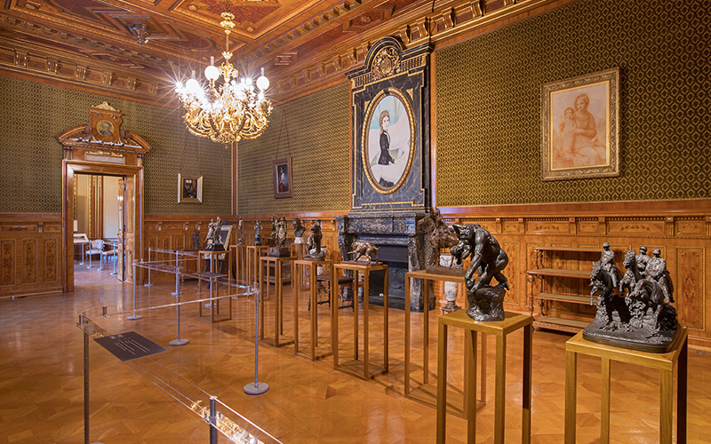 Ein reich verzierter Museumsraum mit goldenen Wänden und Decken, einem Kronleuchter, Skulpturen auf Sockeln und gerahmten Porträts an den Wänden, darunter ein großes Porträt über einem Kamin.