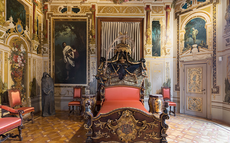 Verziertes, antikes Schlafzimmer mit einem üppigen rot-goldenen Himmelbett, passenden Stühlen, großen klassischen Gemälden und aufwändigem Wanddekor. Eine Statue im Gewand steht in der Ecke auf einem karierten Boden.