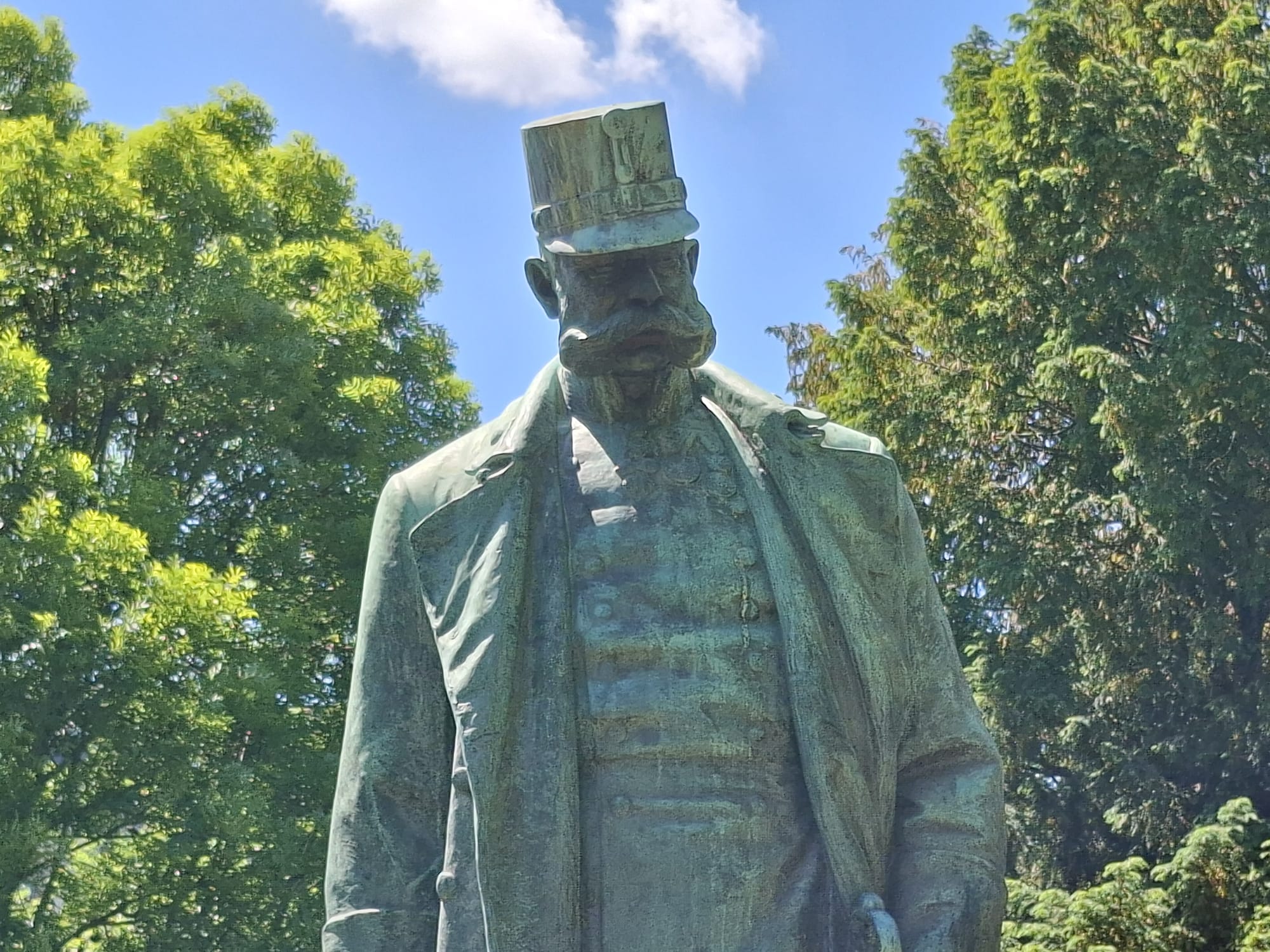 Im Freien steht eine Bronzestatue eines Mannes in Militäruniform und mit Mütze, umgeben von grünen Bäumen und unter einem teilweise bewölkten blauen Himmel.