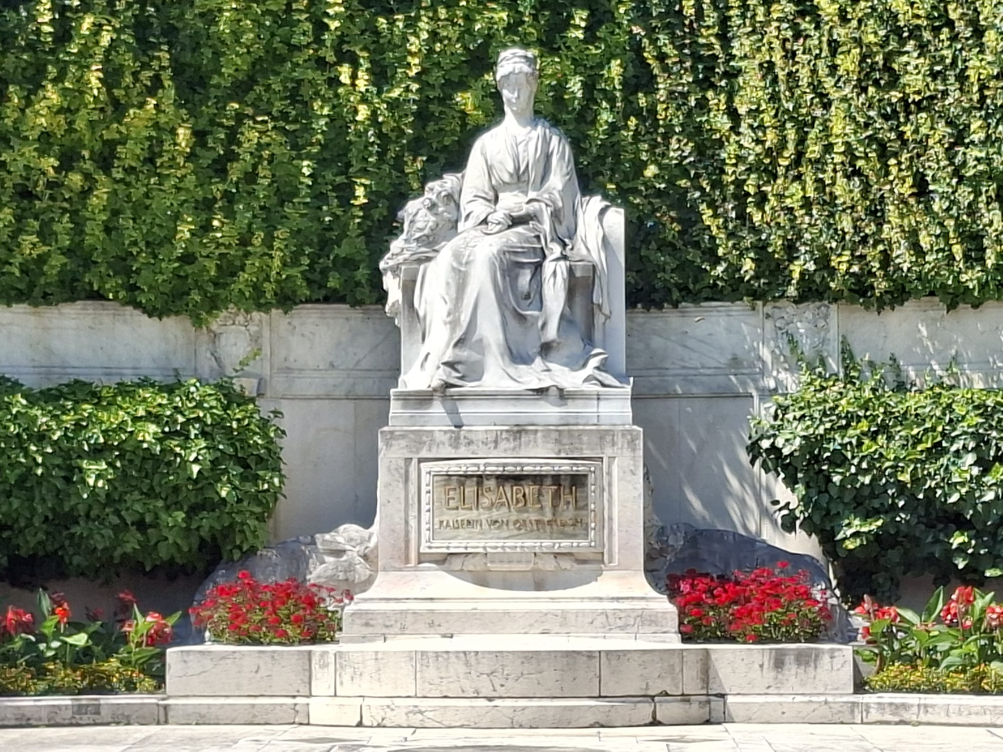 Mramorová socha sedící ženy s lvem po boku, obklopená zelenými rostlinami a červenými květinami. Na podstavci je nápis „Elisabeth“.