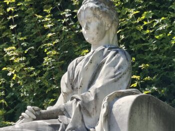 Eine verwitterte Steinstatue einer sitzenden Figur in Gewändern, deren Hände auf dem Schoß ruhen, vor dem Hintergrund grüner Efeublätter. Das Gesicht der Statue ist undeutlich und glatt.